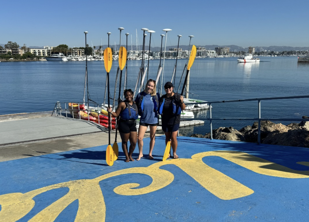 Three young women in life jackets hold paddles, standing on a vibrant blue dock with yellow lettering, overlooking a calm waterway and boats.